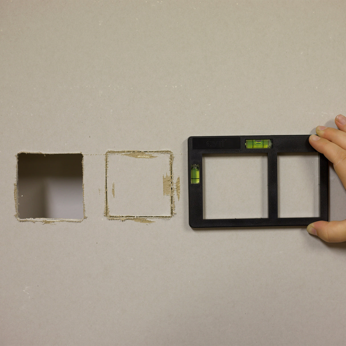 Person demonstrating a AV02034 socket box template with level tool on a wall with one square hole and one scored out hole.